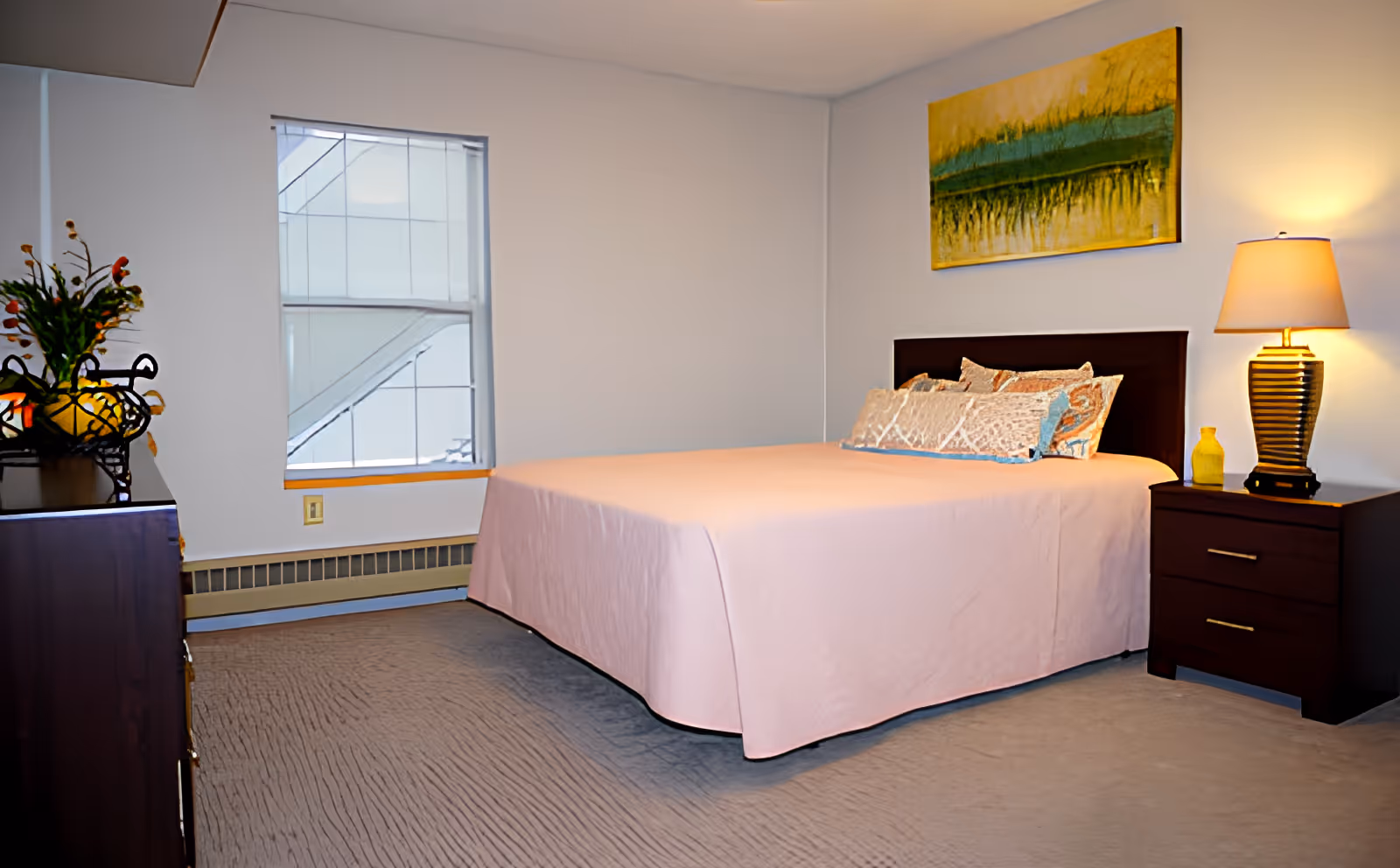 A neatly made bed with a light pink bedspread and decorative pillows in a bedroom. There is a wooden nightstand with a lamp and a small yellow decorative item on it. A colorful abstract painting hangs on the wall above the bed. A window with blinds is on the left side, and a dresser with a flower arrangement is partially visible.