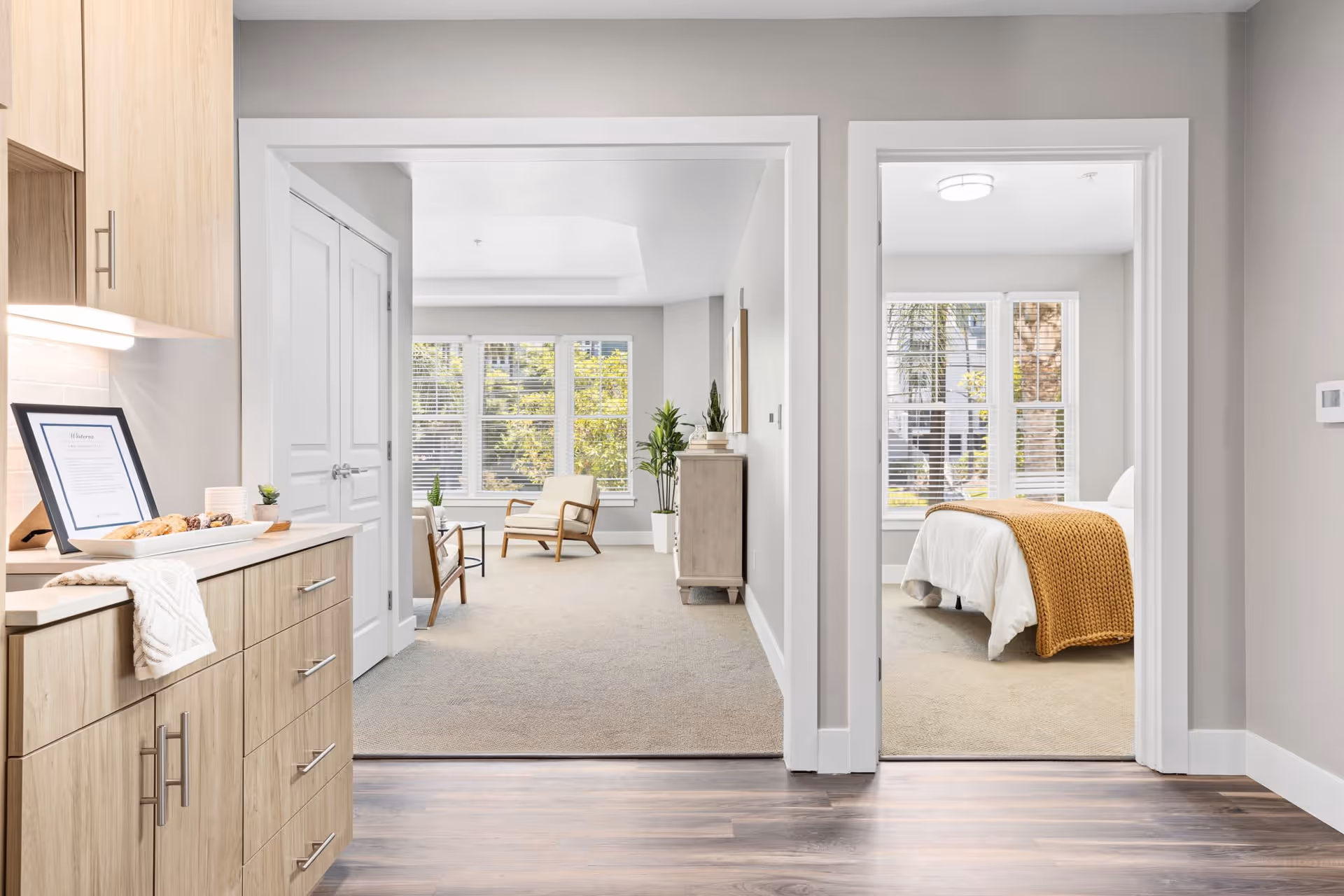 View from a kitchen area into a living room and a bedroom in a senior living facility. The kitchen has light wood cabinets and a countertop with a framed document and some snacks. The living room has beige carpet, two wooden chairs with cushions, a small table, and large windows with blinds letting in natural light. The bedroom has a bed with white bedding and a mustard yellow knitted throw blanket, also with large windows and blinds.