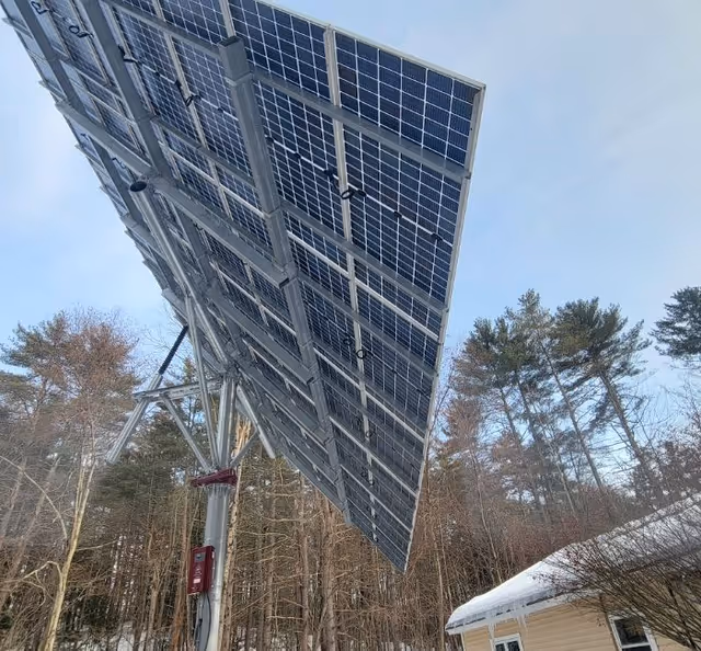 Large solar panel array mounted on a metal structure outdoors with trees and a house with a snow-covered roof in the background under a clear sky.