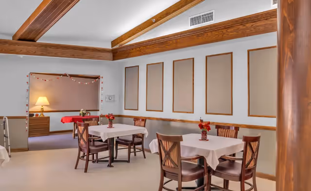 Dining room with small tables covered in white tablecloths, wooden chairs, floral centerpieces, and exposed wooden ceiling beams.