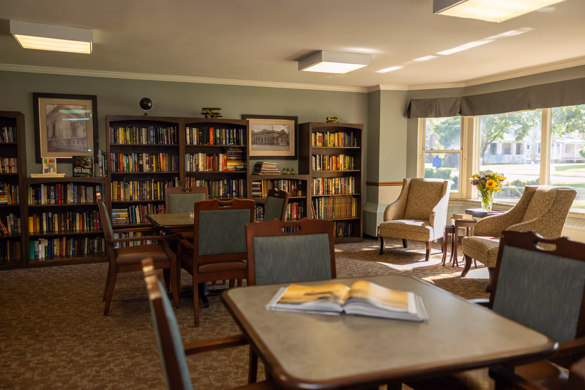 A sunny senior living common room with bookshelves, tables, and armchairs by large windows.