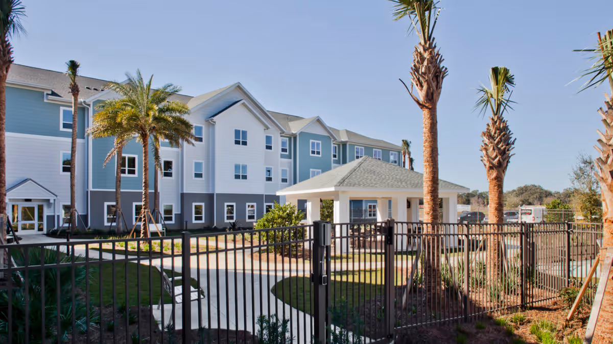 Exterior view of a multi-story senior living facility named Trinity Springs with a fenced garden area featuring palm trees and a covered seating pavilion under a clear blue sky.
