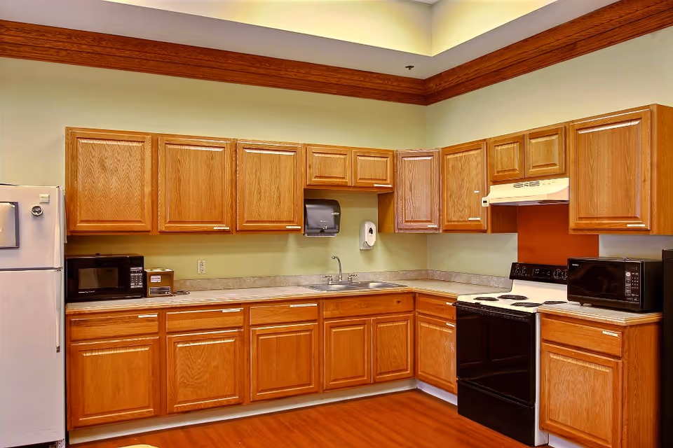 A kitchen area with wooden cabinets, a white refrigerator, two black microwaves, a black stove with an overhead vent, a sink, and a paper towel dispenser mounted on the wall. The walls are painted light green with a brown accent behind the stove, and the floor is wooden.