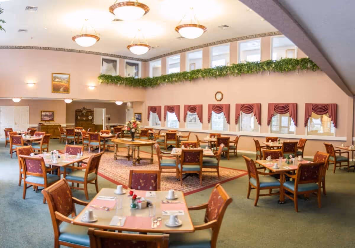A spacious dining room with multiple wooden tables and chairs arranged neatly. Each table is set with cups, saucers, utensils, and small flower vases. The room has large windows with red curtains, green plants along the upper wall, and several ceiling lights providing warm illumination. A round table with a floral centerpiece is placed on a patterned rug in the center of the room.