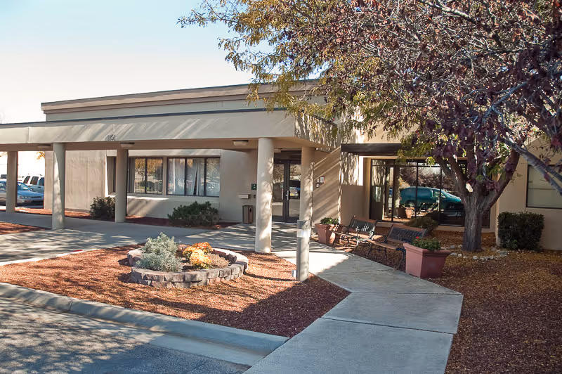 Exterior view of a single-story building entrance with a covered walkway supported by columns. There is a small landscaped area with plants and mulch in front, a concrete sidewalk leading to the entrance, benches, and a tree with autumn foliage nearby.