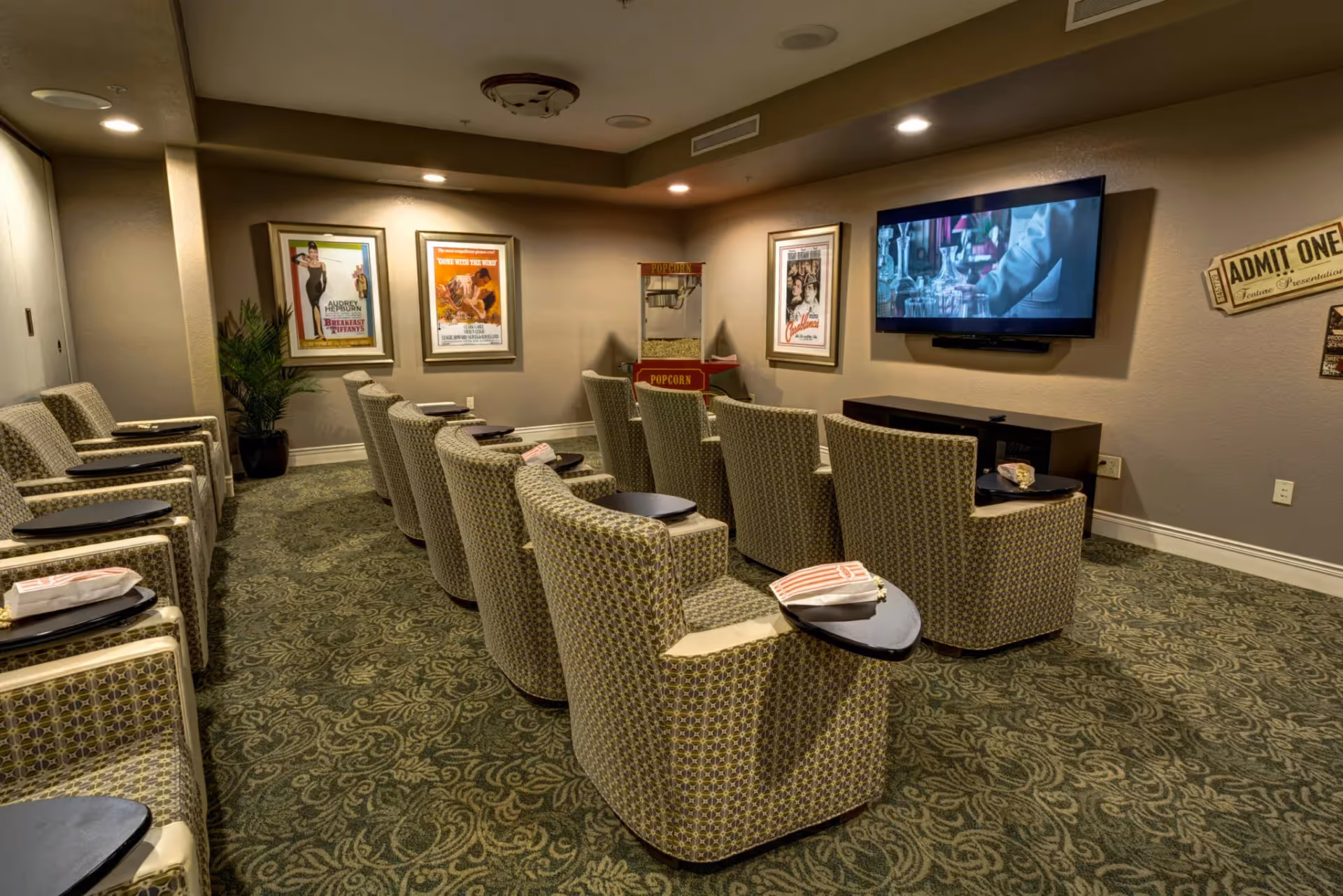 A small movie theater room with two rows of patterned armchairs, each equipped with a small attached table holding popcorn bags. The room has a green patterned carpet, framed vintage movie posters on the walls, a popcorn machine in the corner, and a flat-screen TV mounted on the wall showing a scene from a movie.