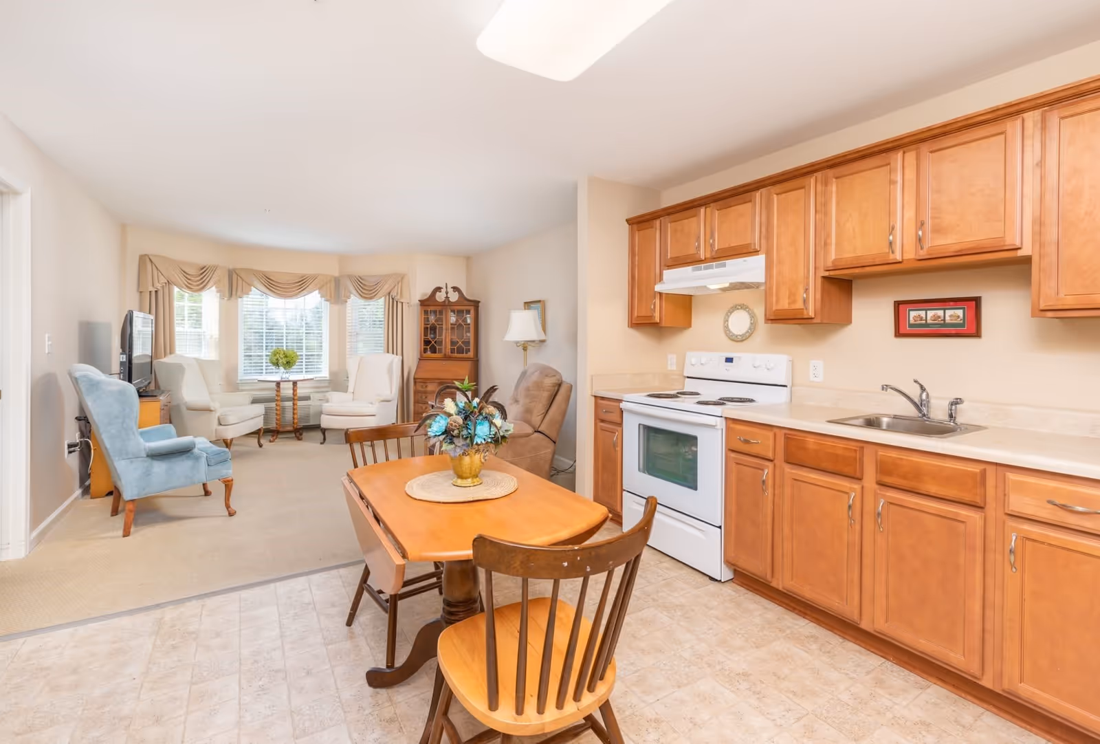Bright open-plan kitchen and sitting area with wooden cabinets, a small dining table, and upholstered chairs by a bay window.