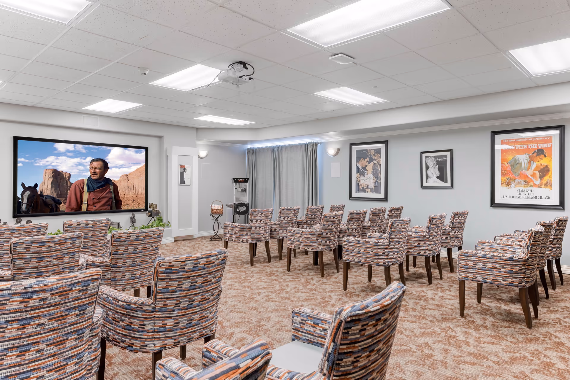A senior living facility media room with multiple rows of patterned upholstered chairs facing a large screen showing a western movie scene. The room has beige carpet, light gray walls, framed classic movie posters, and a popcorn machine in the corner.