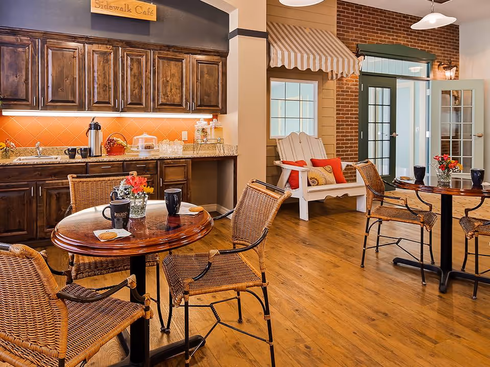 Cozy café area inside a senior living facility with wooden tables and wicker chairs. A countertop with a coffee station and snacks is visible against the wall with wooden cabinets above. There is a white Adirondack chair with red and beige cushions near a window and a door with glass panels. The floor is wooden, and the walls feature a mix of brick and painted surfaces. A small wooden sign above the cabinets reads 'Sidewalk Café'.