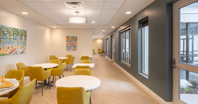 Bright communal dining area with round white tables, chartreuse upholstered chairs, artwork on the wall, and windows along the right side.