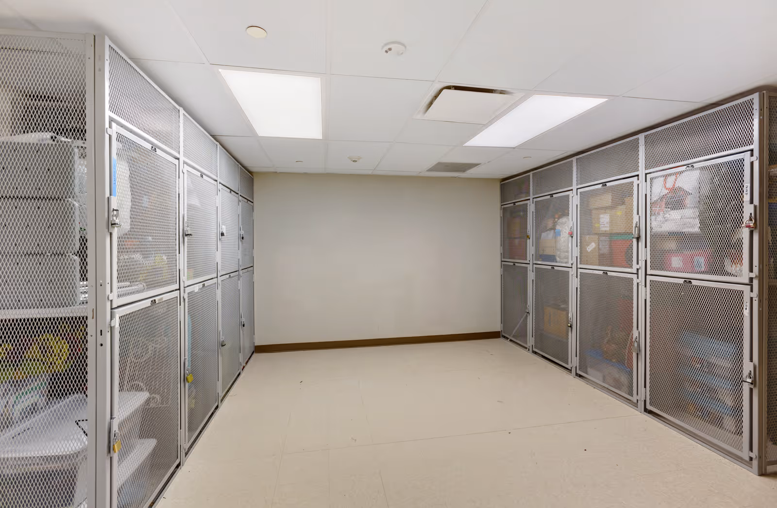 A storage room with metal mesh lockers on both sides filled with various boxes and items. The room has a tiled floor, a plain beige wall at the far end, and a white drop ceiling with fluorescent lighting panels.