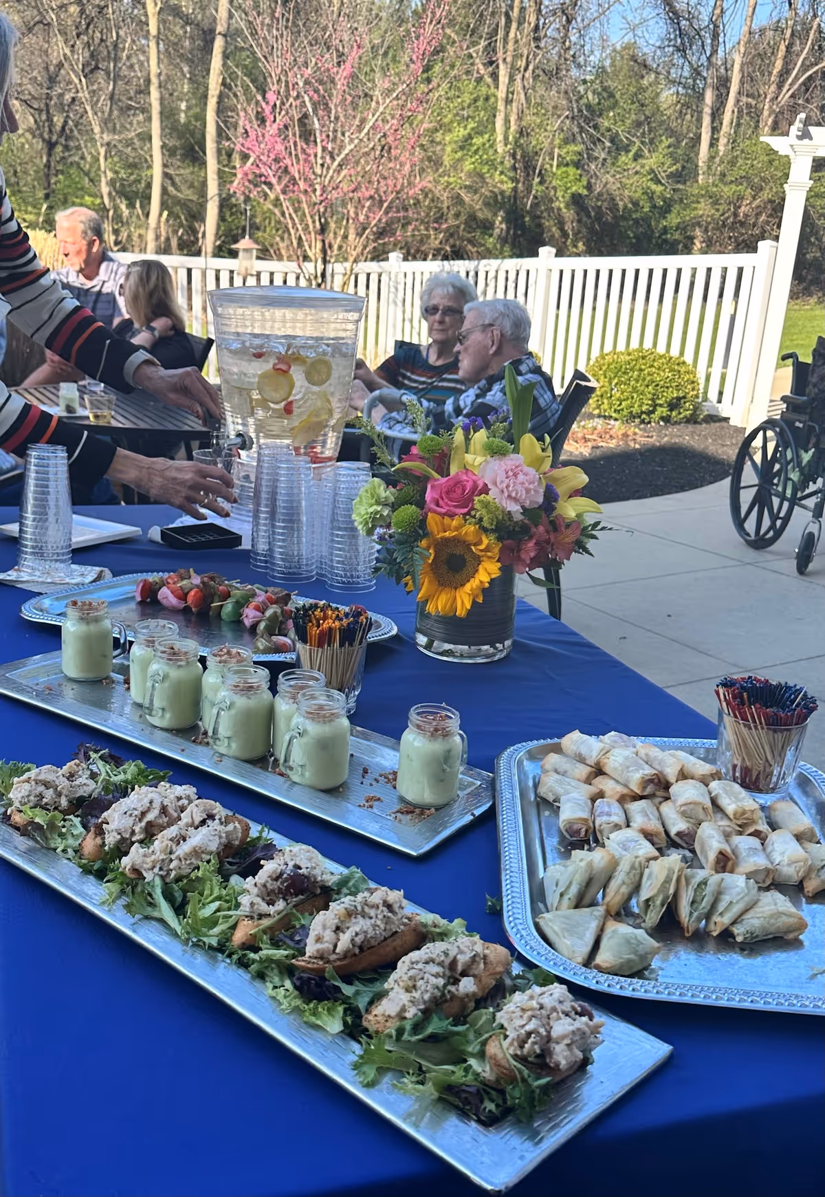 Outdoor gathering at Arbor Terrace Willistown with a table covered in a blue tablecloth displaying trays of appetizers including small sandwiches, spring rolls, and jars of a creamy dessert. A large beverage dispenser with fruit-infused water and stacks of clear plastic cups are also on the table. In the background, elderly people are seated and conversing near a white fence with trees and greenery behind them. A colorful flower arrangement with sunflowers and roses is placed on the table.