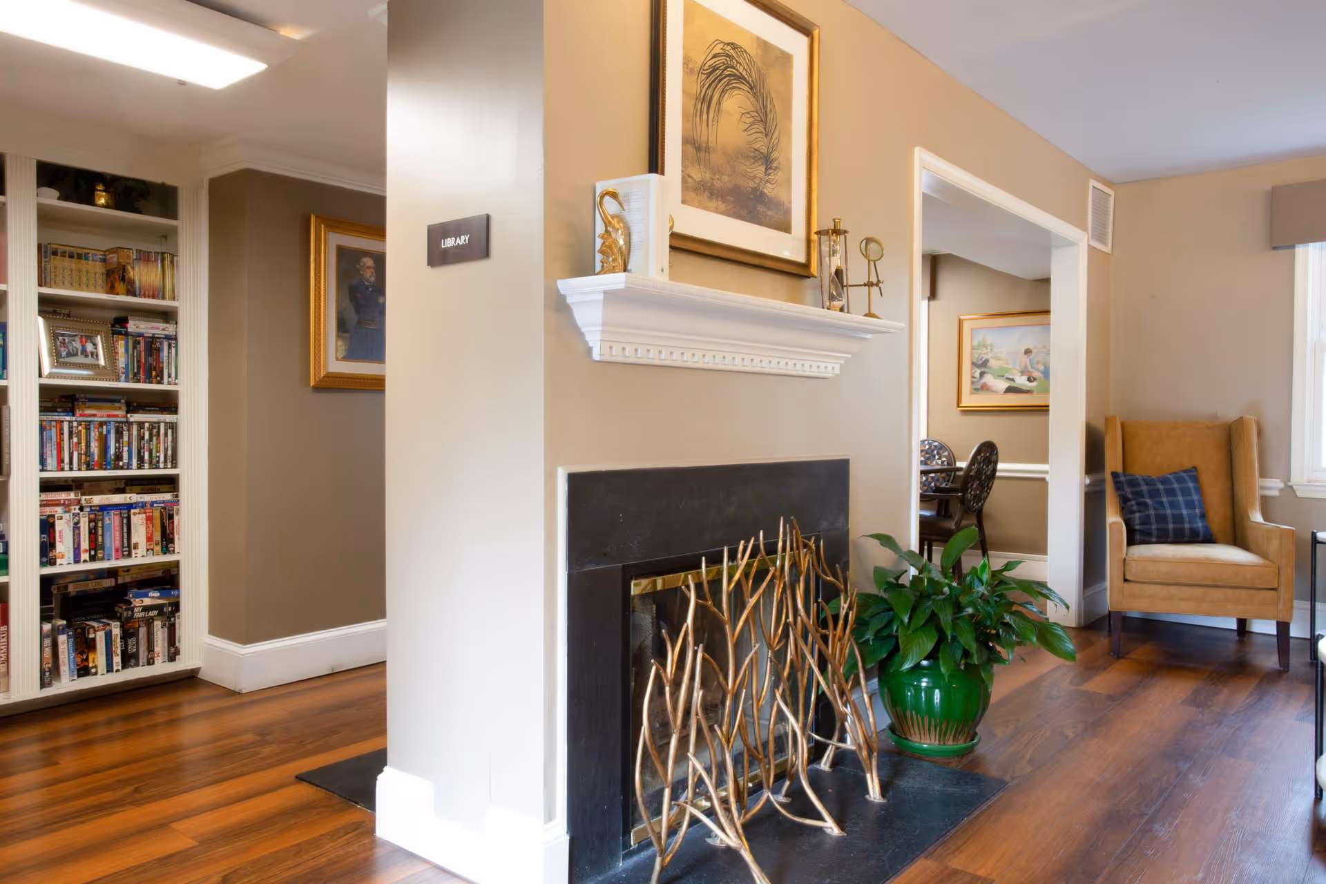 Cozy interior space featuring a fireplace with decorative golden branches in front, a green potted plant beside it, and a beige armchair with a blue plaid pillow near a window. To the left, there is a built-in bookshelf filled with books and DVDs, and a sign on the wall indicating the library. The room has warm wooden flooring and neutral-colored walls with framed artwork.