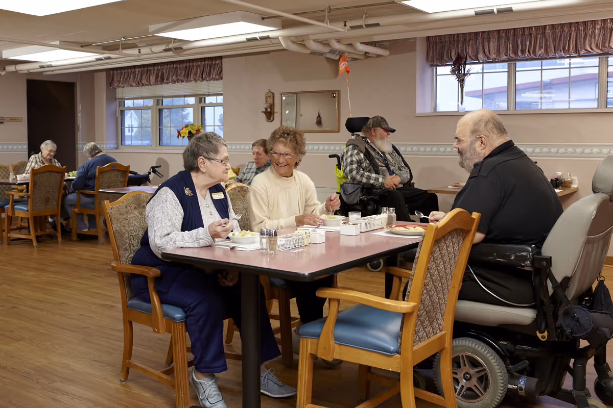 Several elderly individuals sitting and eating together in a dining room at an assisted living facility. The room has wooden floors, large windows with curtains, and tables with chairs. One man is in a motorized wheelchair, and others are engaged in conversation while enjoying their meals.