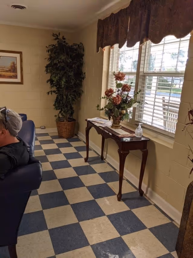 Interior corner of a room with checkered blue and white tile floor, a tall potted plant in the corner, a small wooden table with a floral arrangement and hand sanitizer on it, and a window with blinds and a valance. A person wearing a cap is partially visible sitting on a blue chair on the left side.