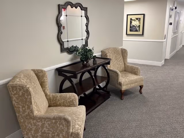 A hallway seating area in a senior living facility featuring two beige patterned armchairs positioned on either side of a dark wooden console table with a decorative plant on top. Above the table is a decorative mirror with a scalloped frame. The hallway has light-colored walls, carpeted flooring, and a framed artwork on the wall further down the corridor.