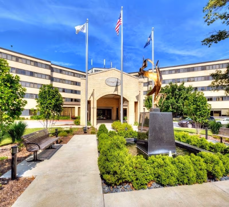 Exterior view of a multi-story senior living facility with a central entrance featuring an archway. In front of the entrance is a landscaped area with a water fountain and a modern bronze sculpture. Three flagpoles with flags stand behind the fountain. Benches and greenery line the walkway leading to the entrance under a clear blue sky.