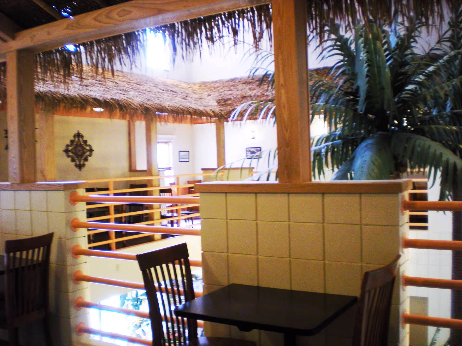 Indoor seating area with wooden chairs and tables, surrounded by a railing and a partial thatched roof structure. There are large artificial palm trees and decorative wall art in the background, creating a tropical-themed interior space.