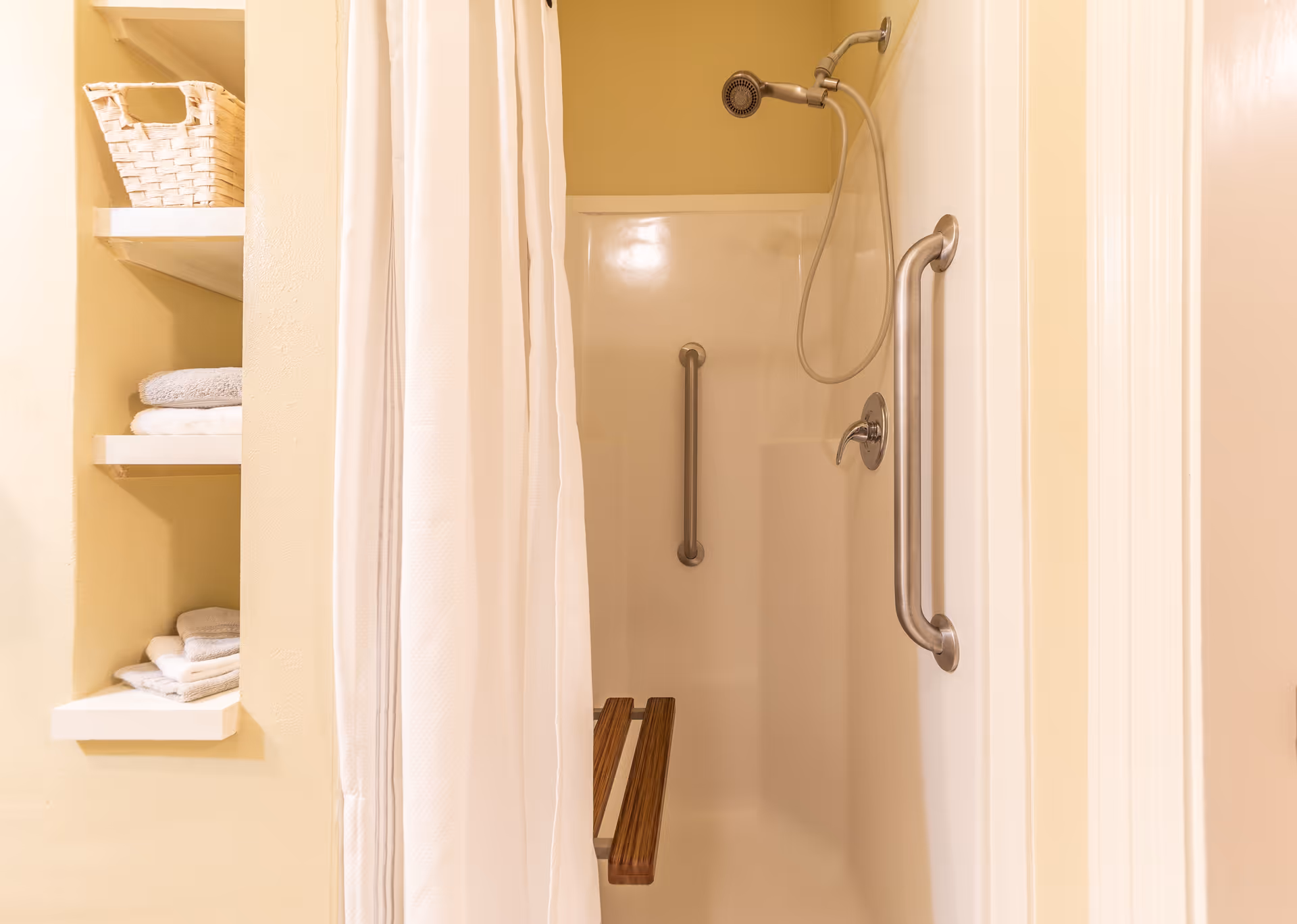 A shower area with a white curtain partially drawn to the side, featuring a wall-mounted showerhead, two metal grab bars, and a foldable wooden shower seat. To the left, there are built-in shelves holding neatly folded towels and a wicker basket.