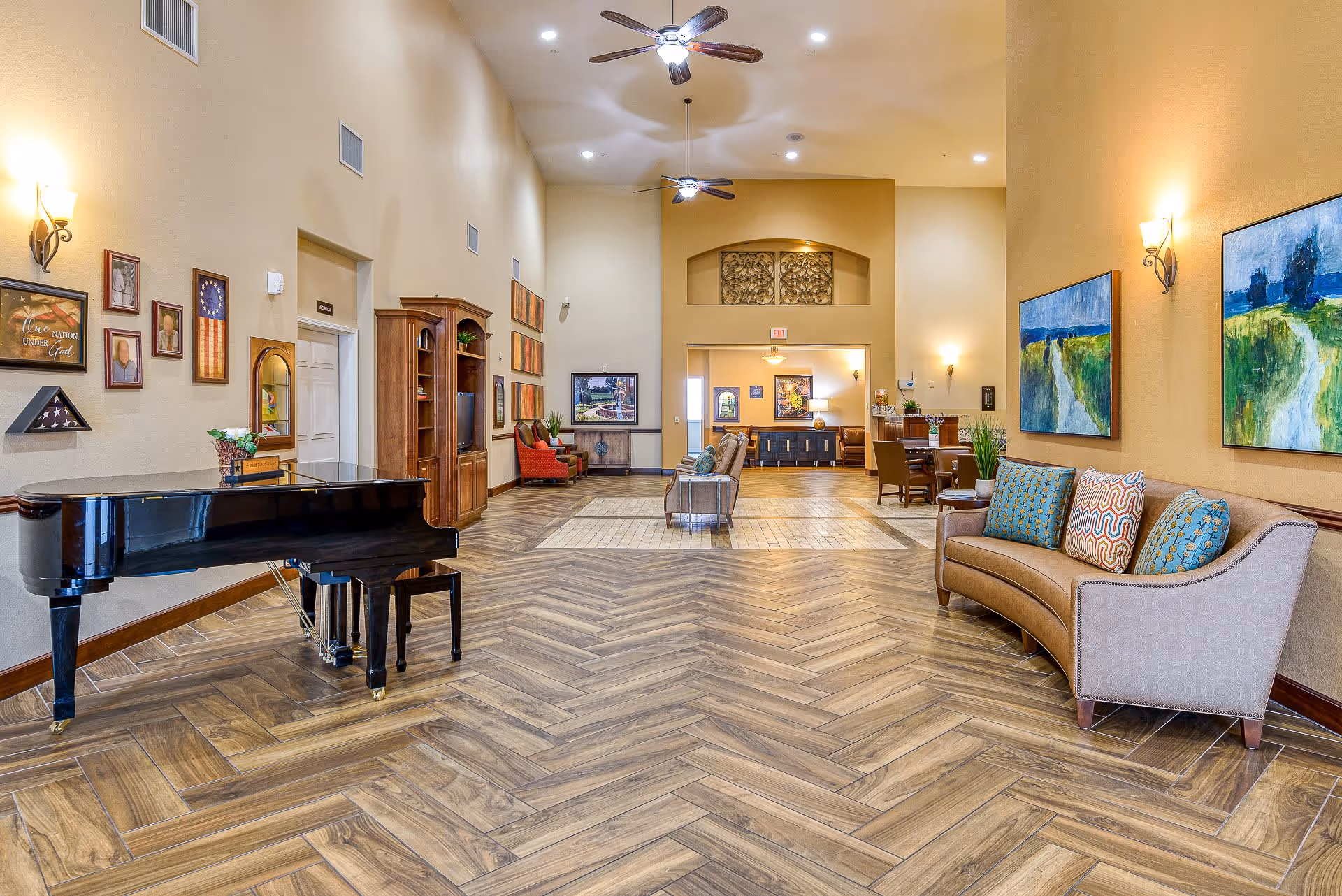 A spacious senior living facility common area with wooden herringbone patterned flooring, beige walls, and high ceilings with ceiling fans. The room features a black grand piano on the left, a curved beige sofa with colorful pillows on the right, several framed paintings and photographs on the walls, and various seating areas with chairs and tables in the background.