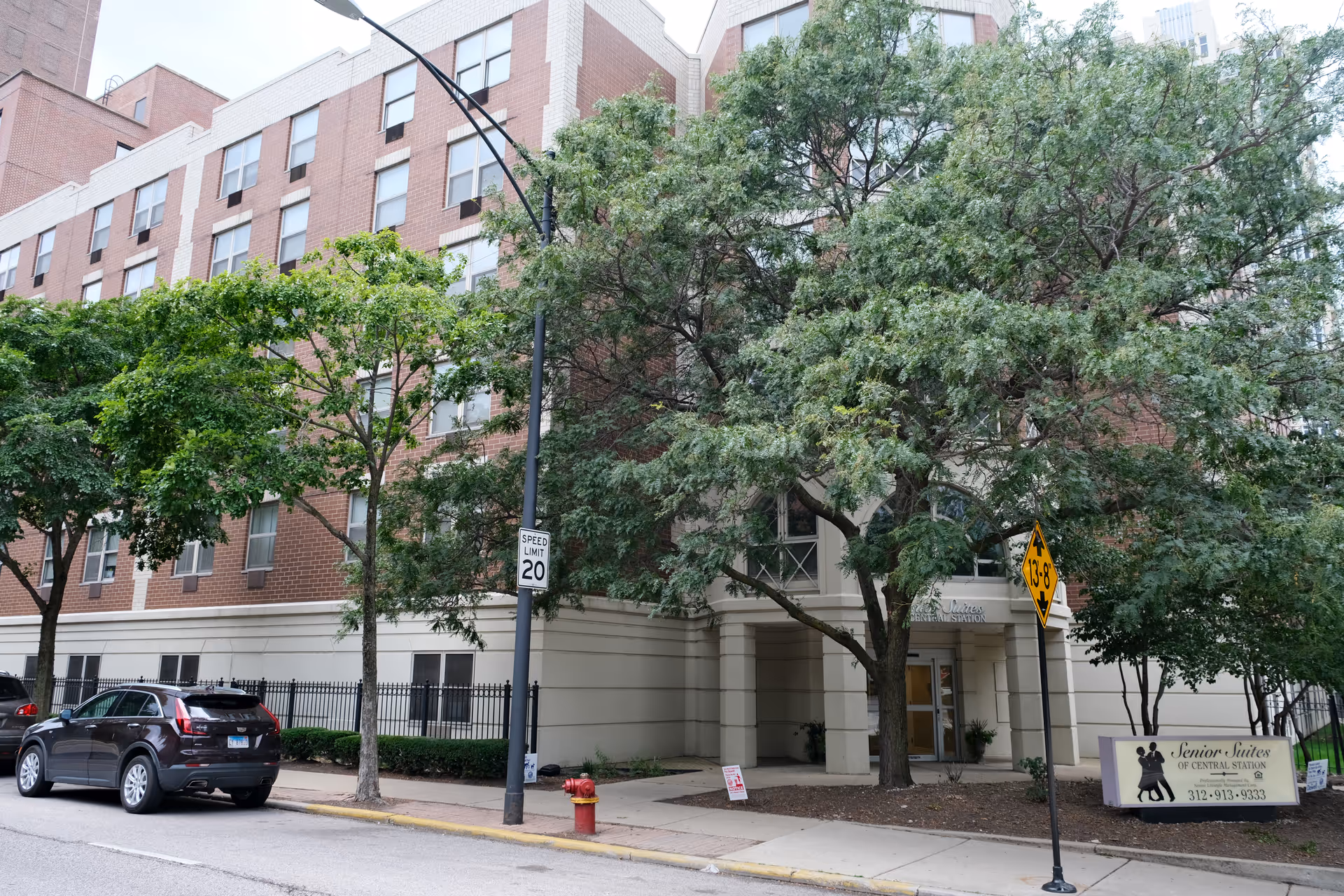 Exterior view of Senior Suites of Central Station, a multi-story brick and beige building partially obscured by trees. A black car is parked on the street in front of the building, and there are street signs including a speed limit sign and a height clearance sign. A sign near the entrance displays the facility name and phone number.