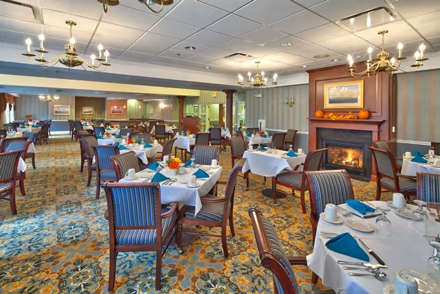 A spacious dining room with multiple tables covered in white tablecloths, each set with plates, cups, silverware, and teal napkins. The room features patterned carpet, striped wallpaper, chandeliers, and a lit fireplace with a framed picture above it. The atmosphere is warm and inviting.