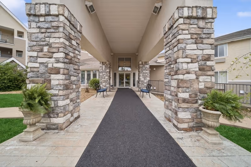 Covered entrance walkway to a senior living building with stone pillars, benches, and potted ferns leading to glass doors.
