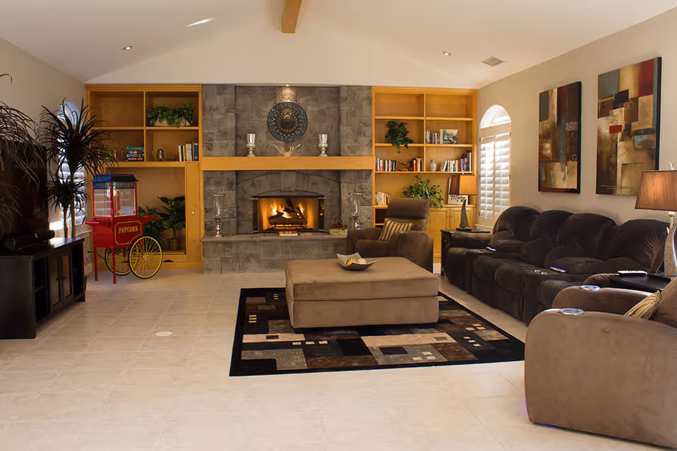 Spacious living room with a stone fireplace flanked by built-in shelves, plush reclining sofas, a central ottoman on a patterned rug, and a popcorn cart.