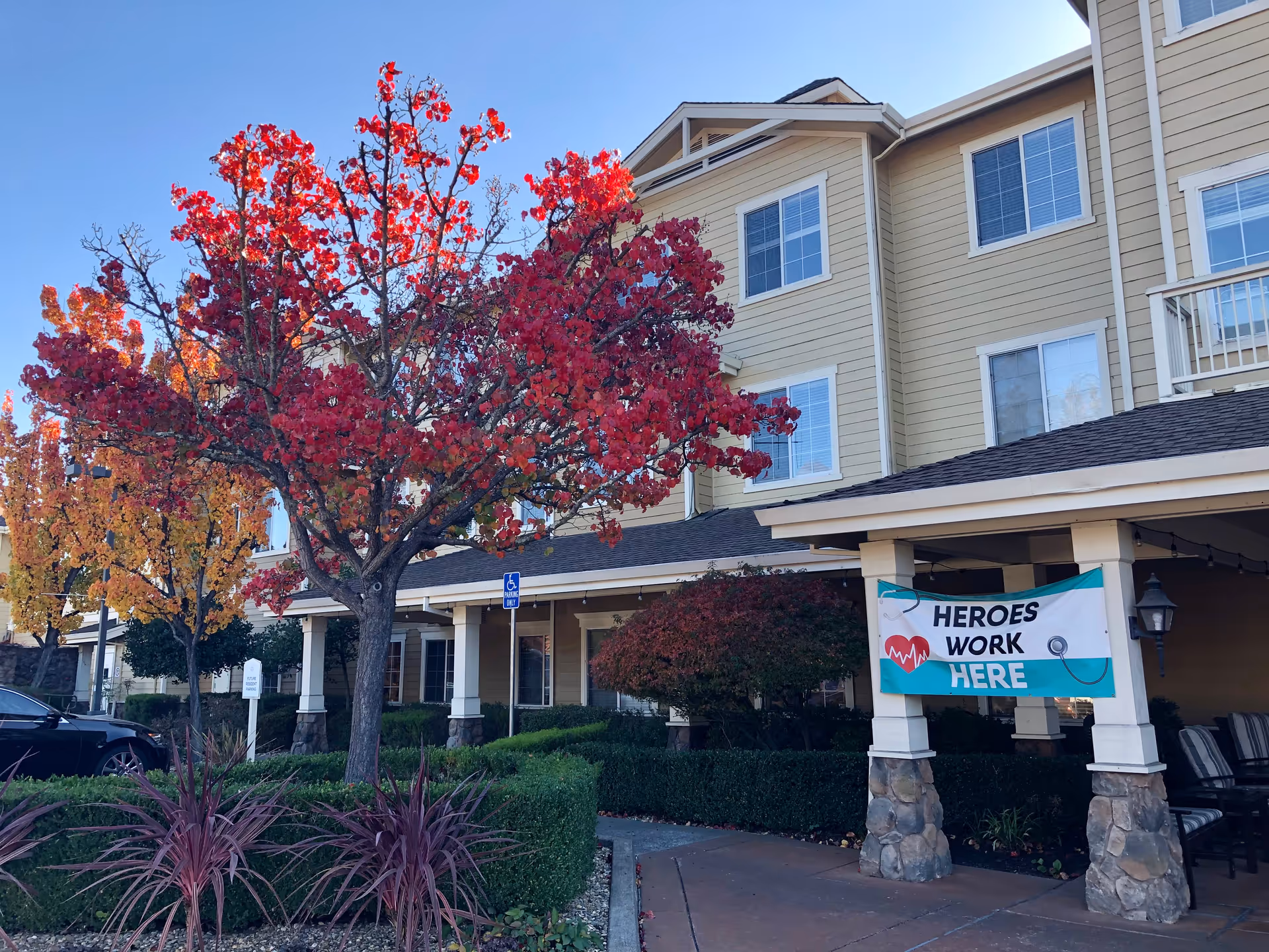 Exterior view of Arbol Residences of Santa Rosa building with beige siding and multiple windows. In front, there are trees with red and orange autumn leaves, bushes, and a sidewalk. A banner hanging on the porch reads 'HEROES WORK HERE' with a heart and stethoscope graphic.