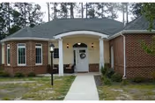 Single-story brick building entrance with a white arched portico, front door with a wreath, a walkway and lamppost flanked by grass and trees.