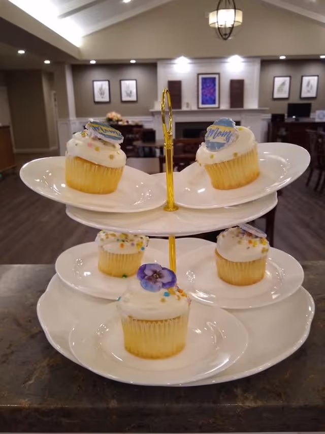 A three-tiered white serving tray with five vanilla cupcakes topped with white frosting and colorful sprinkles. Two cupcakes on the top tier have decorative toppers that say 'Mom'. The background shows a cozy interior with a fireplace, framed artwork, and wooden furniture in a senior living facility.