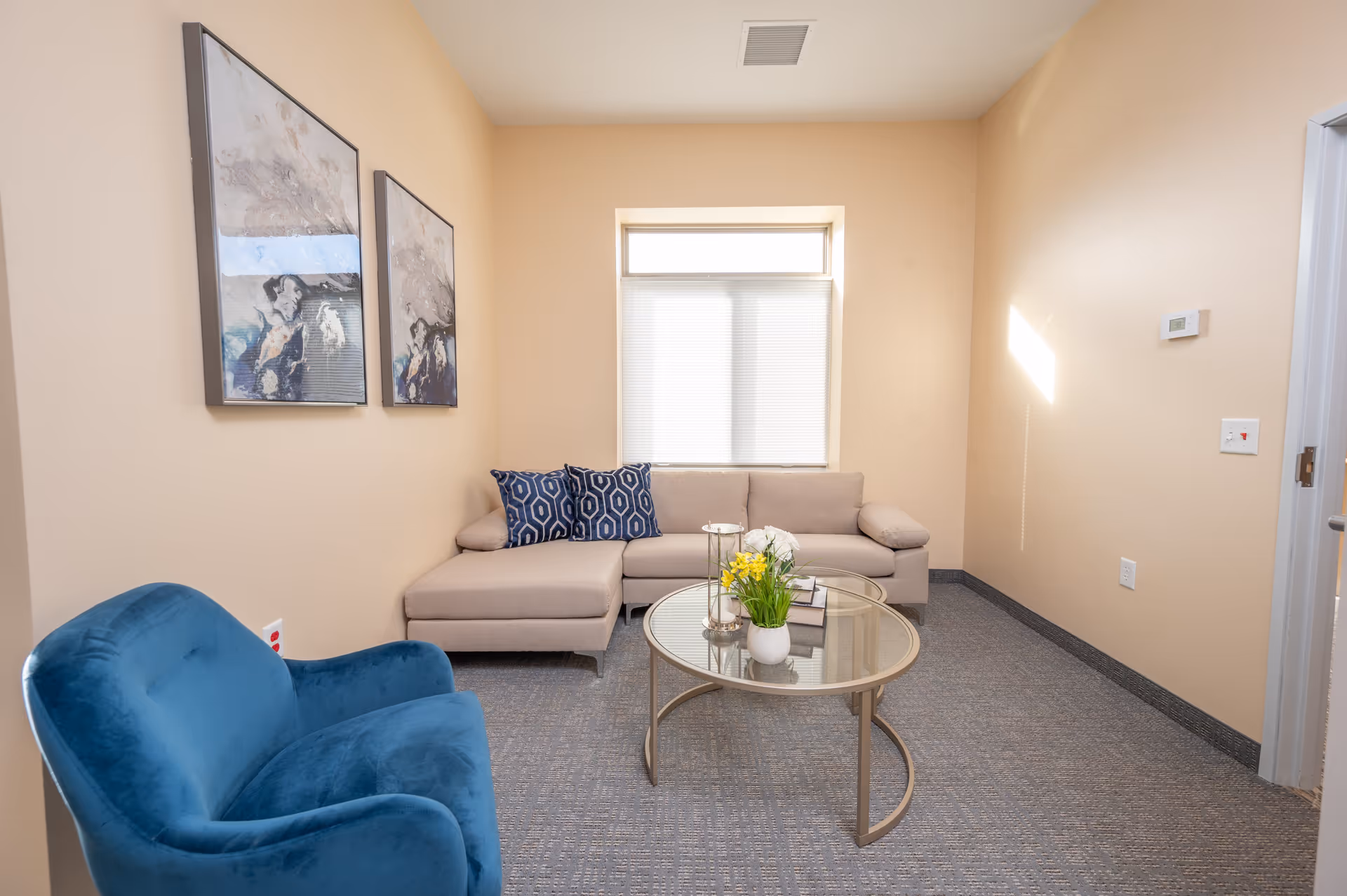 Small sitting room with a beige sectional sofa, blue armchair, round glass coffee table, and framed artwork under a window.