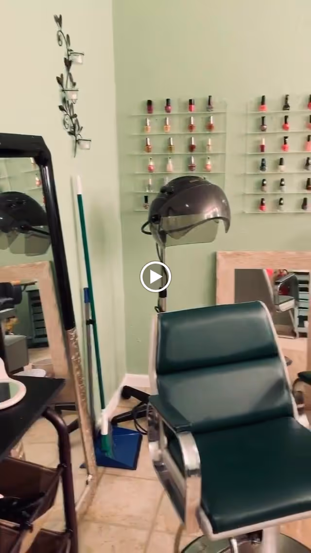 Interior of a salon area with a green salon chair, a hair dryer hood, a wall-mounted rack displaying various nail polish bottles, a large mirror, and cleaning tools like a broom and dustpan against a light green wall.