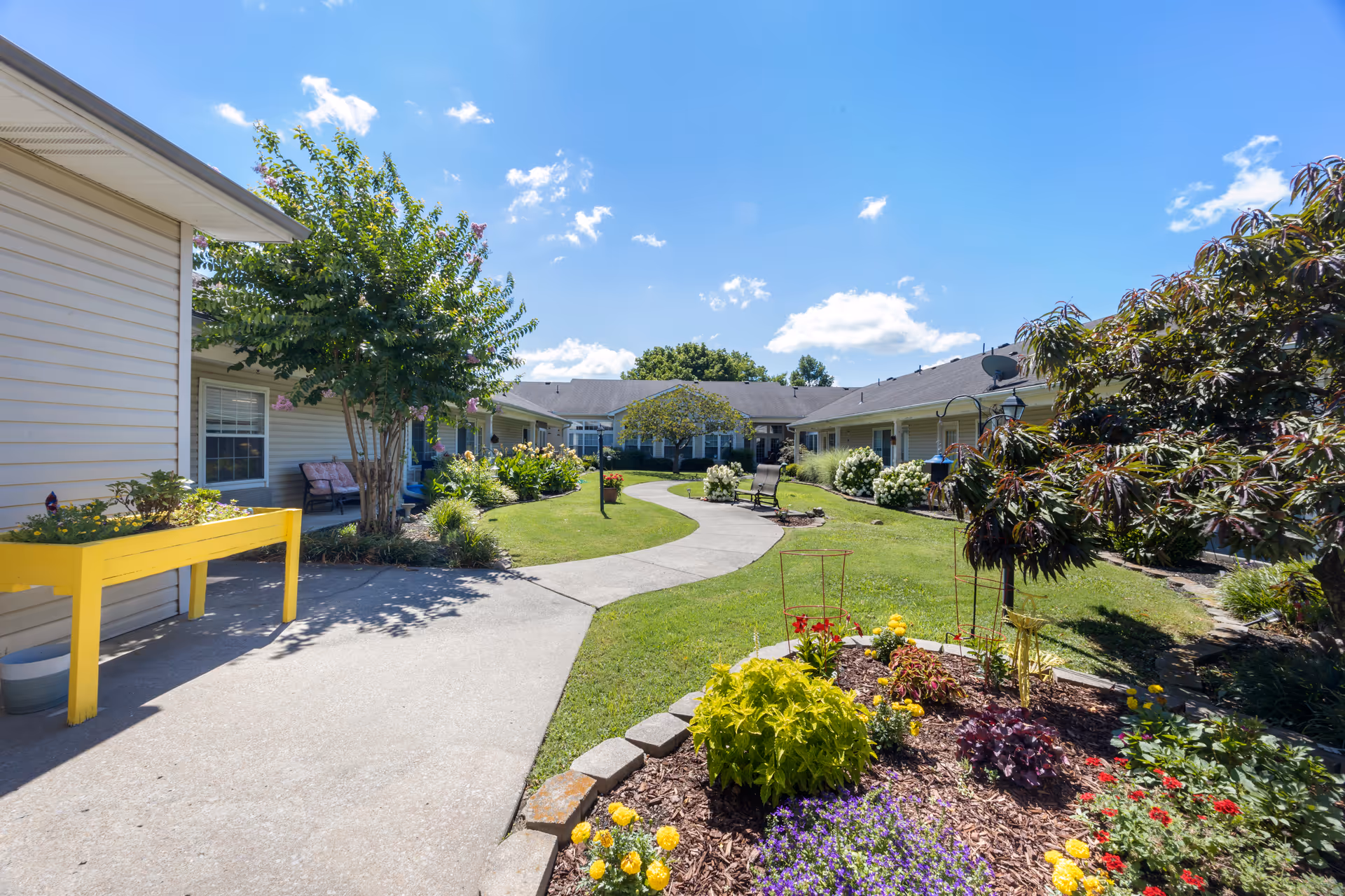 A sunny outdoor courtyard area at a senior living facility with a winding concrete pathway, green grass, colorful flower beds, and trees. The single-story building with beige siding surrounds the courtyard, and there are benches and a yellow raised garden bed along the path.