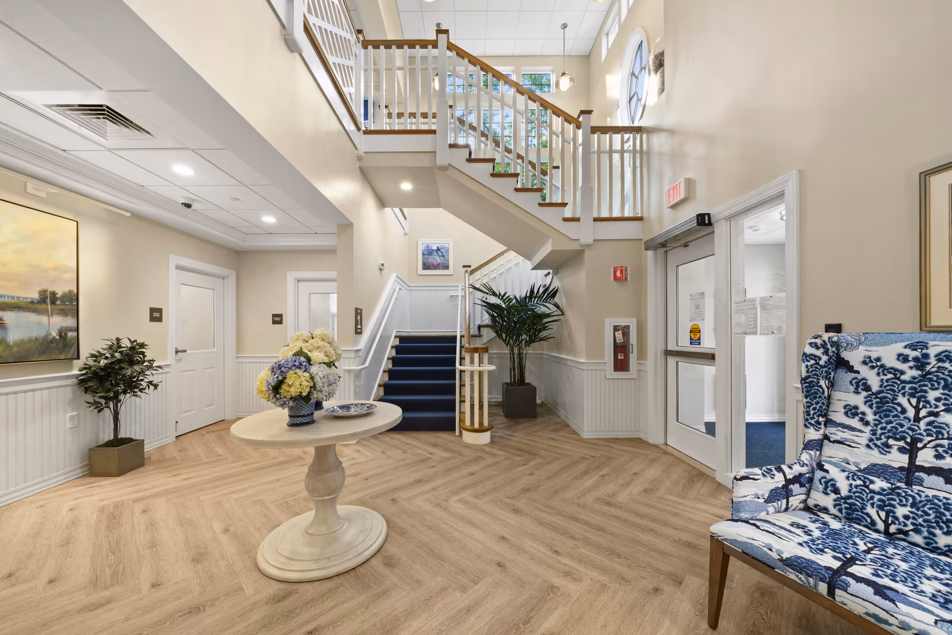 Bright and spacious interior hallway of a senior living facility with a central round table holding a vase of flowers, a staircase with white railings and wooden handrails leading to an upper floor, potted plants, a blue and white patterned armchair, and doors leading to other rooms.