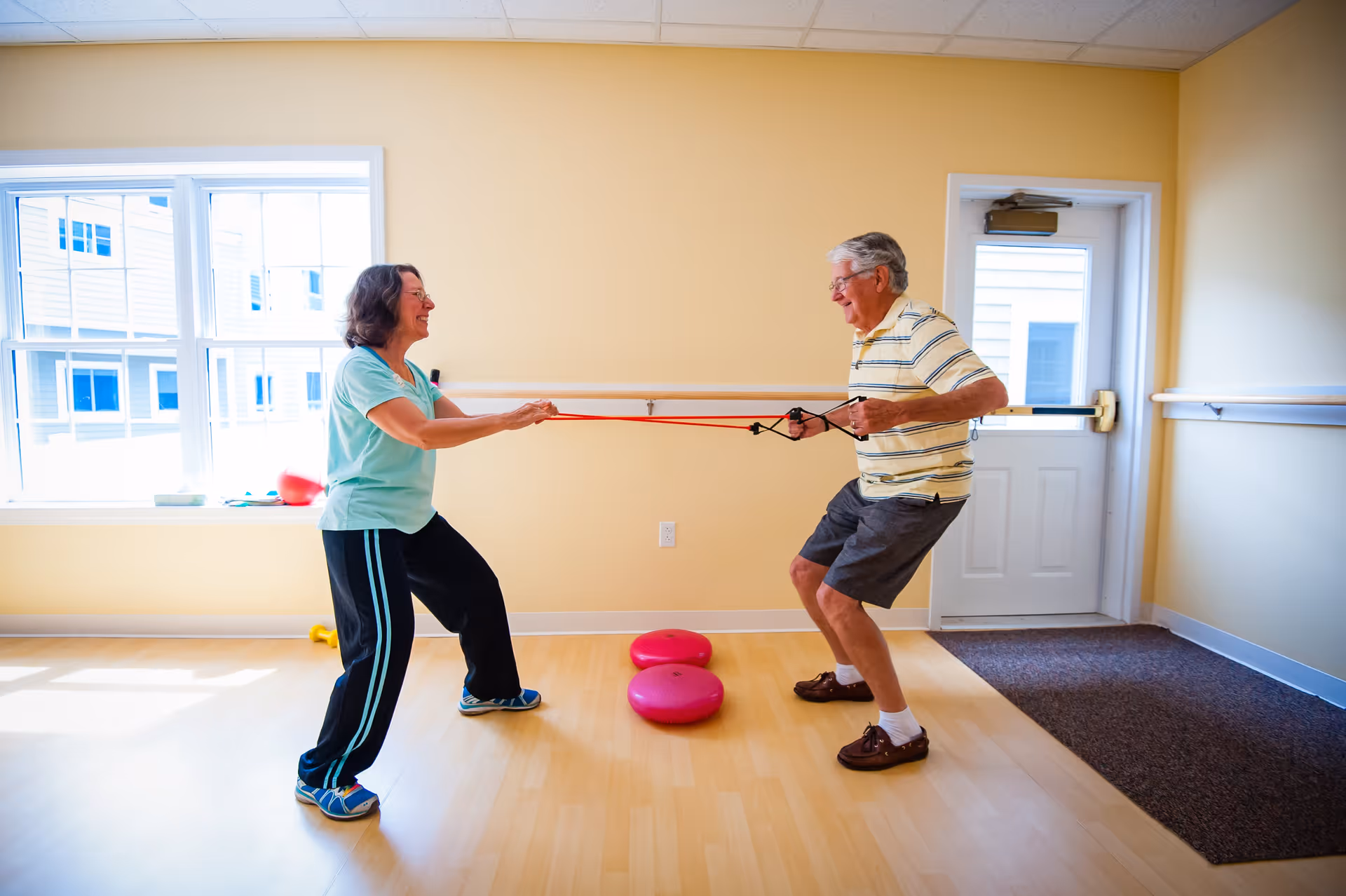 An elderly man and woman are exercising indoors using a resistance band. They are facing each other, pulling the band in opposite directions, smiling and engaged in the activity. The room has light yellow walls, a large window, a door, and wooden flooring with some exercise equipment on the floor.