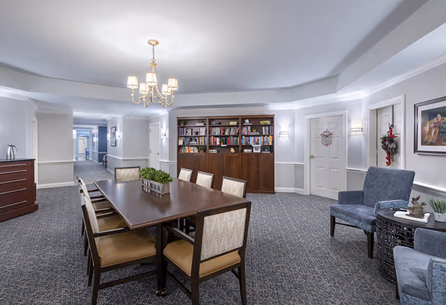 A well-lit interior room with a long wooden table surrounded by eight chairs. There is a chandelier hanging above the table. In the background, there is a large bookshelf filled with books and two closed doors. To the right, there are two blue upholstered chairs with a small round table between them, decorated with a plant and a small sculpture. The room has carpeted flooring and light-colored walls with wainscoting.
