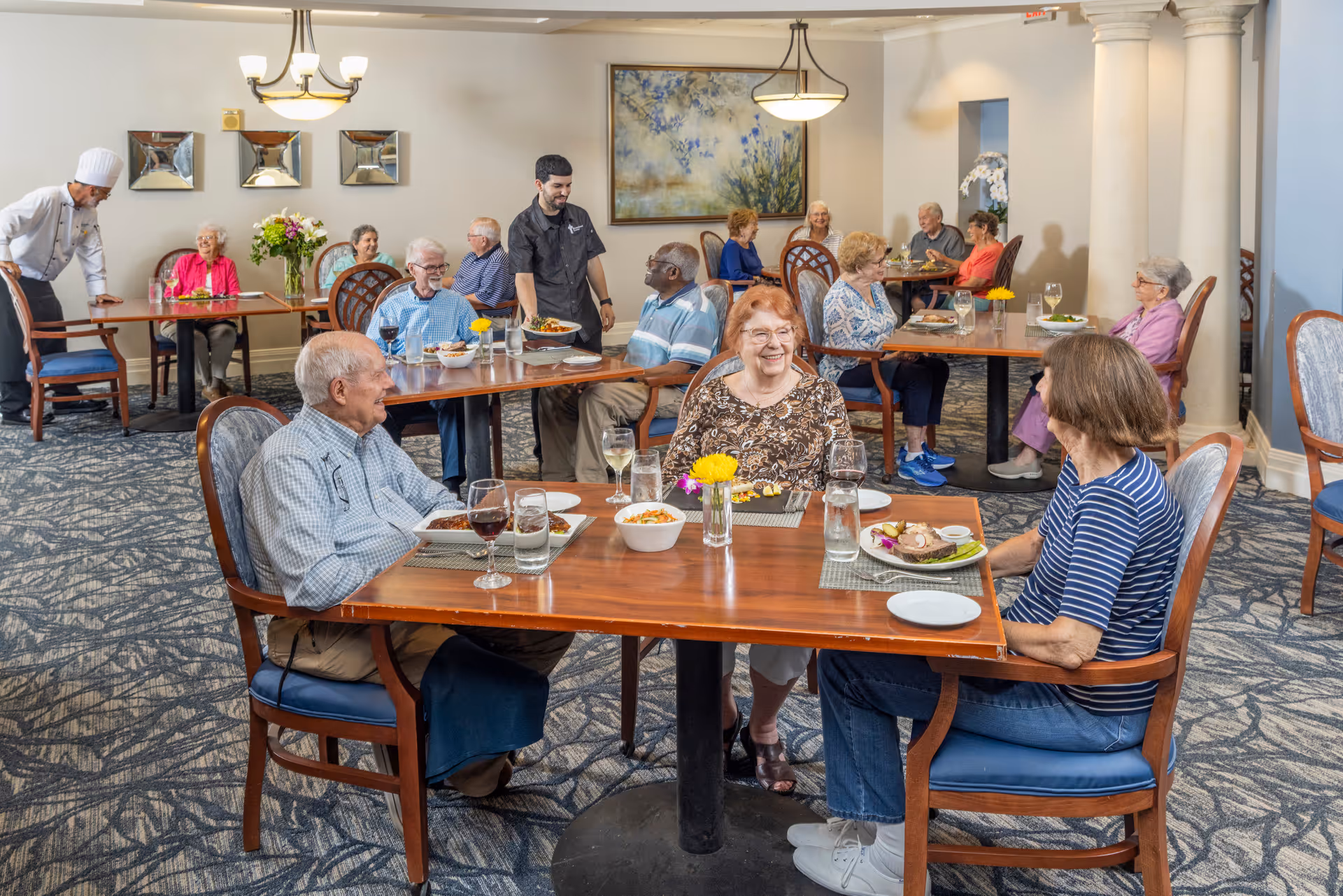 A dining room in Westminster Towers with elderly residents seated at tables enjoying meals. A waiter is serving food, and a chef is standing near the back. The room is well-lit with chandeliers, decorated with flowers and artwork, and has a patterned carpet.