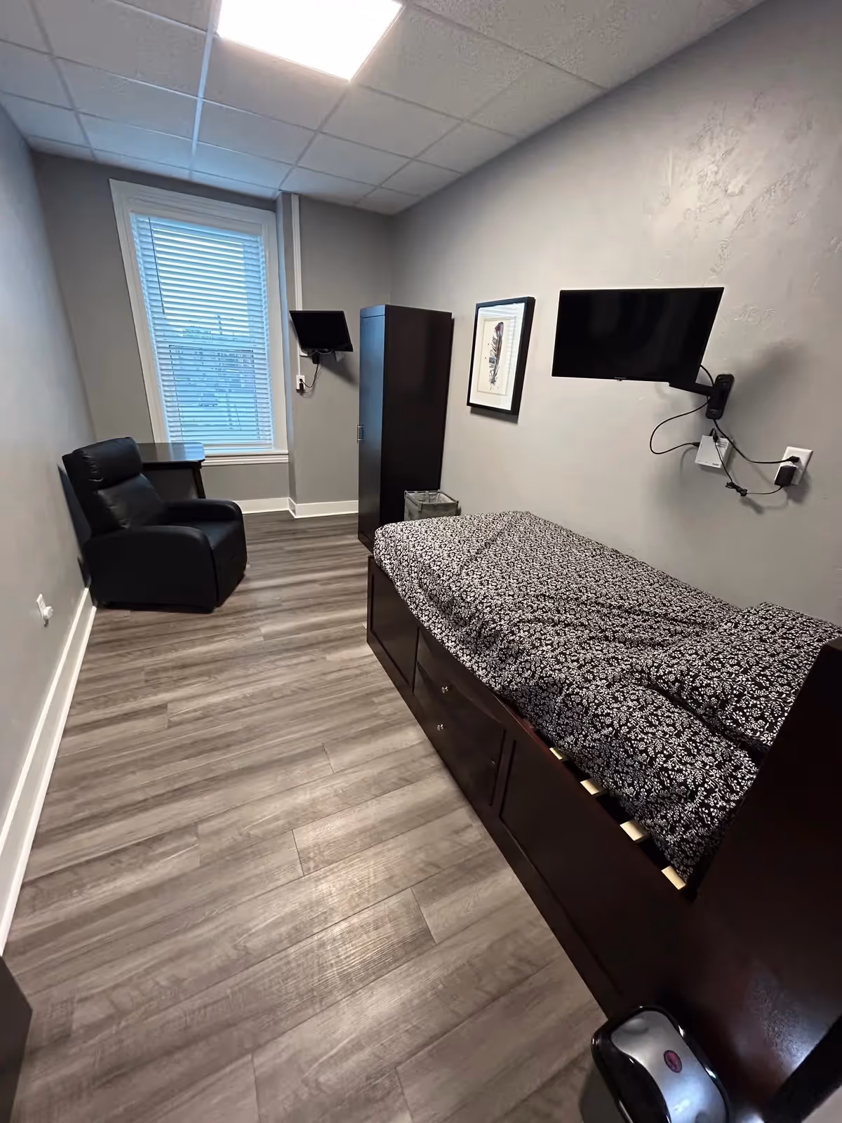 A small, simple bedroom with a single bed covered in a black and white patterned blanket. The room has light gray walls and wood-look flooring. There is a black armchair and a small table near a window with blinds. A dark wooden wardrobe stands against the wall, and a flat-screen TV is mounted on the wall above the bed. A framed picture hangs next to the TV.