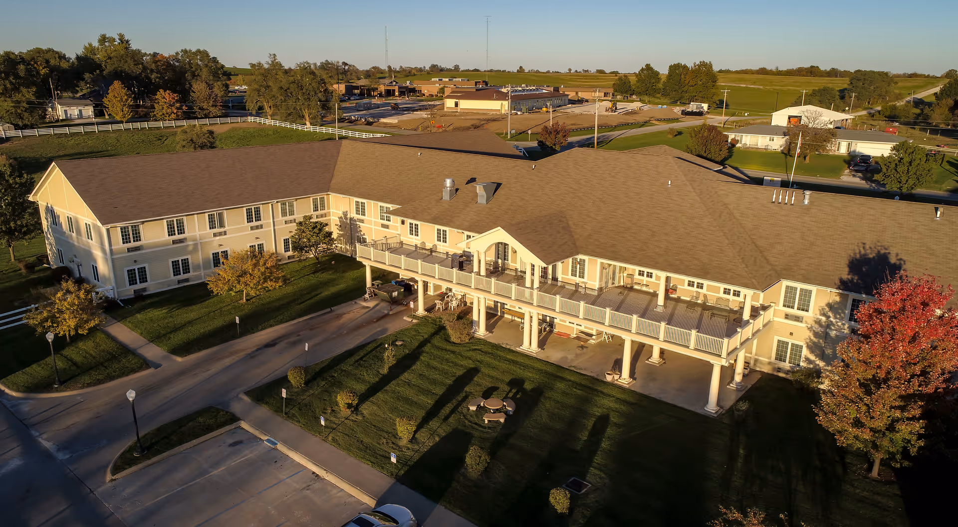 Aerial view of a large two-story assisted living facility building with a beige exterior and a brown roof, surrounded by green lawns, trees, and a parking lot. The building has a covered porch area with white columns and outdoor seating. The surrounding area includes open fields and some other buildings in the distance under a clear blue sky.