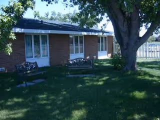 Outdoor area of a brick building with several windows and doors, featuring two benches on a grassy lawn under a large tree with a white fence in the background.