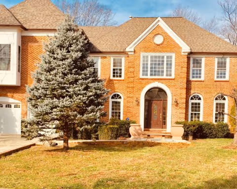 Front exterior of a red-brick two-story building with a central arched entrance, large windows, a front lawn, and a tree.