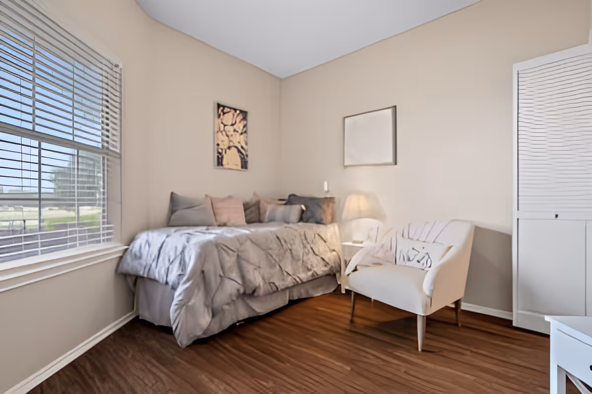 A cozy bedroom with a bed covered in a gray comforter and multiple pillows, a white armchair with a blanket draped over it, a small side table with a lamp, two framed pictures on the beige walls, a window with blinds letting in natural light, and a white closet door.