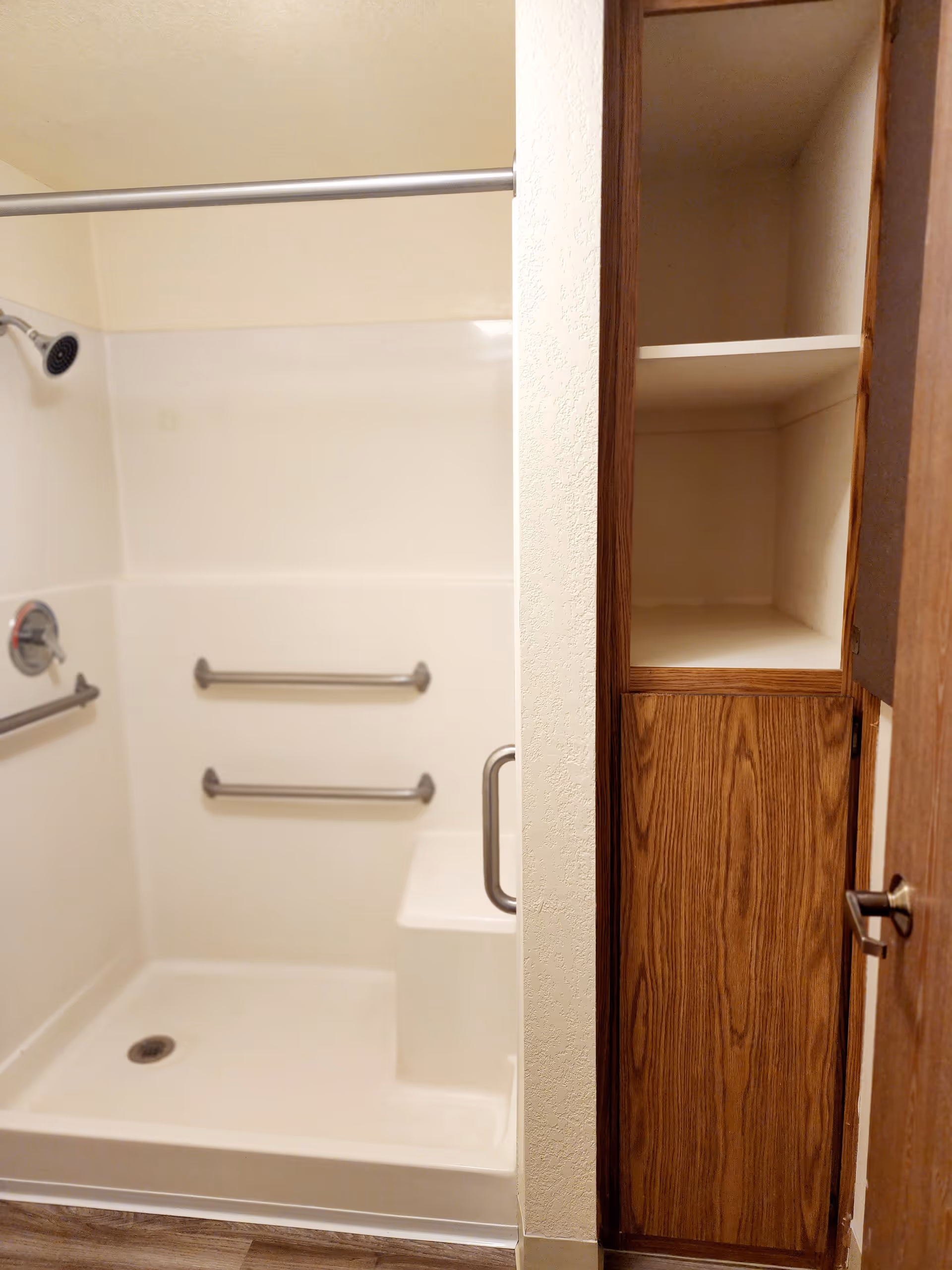 A bathroom shower area with white walls and built-in seat. There are two horizontal grab bars on the wall and a vertical grab bar near the seat. A showerhead is mounted on the left wall. To the right of the shower is a wooden cabinet with open shelves and a closed compartment below.