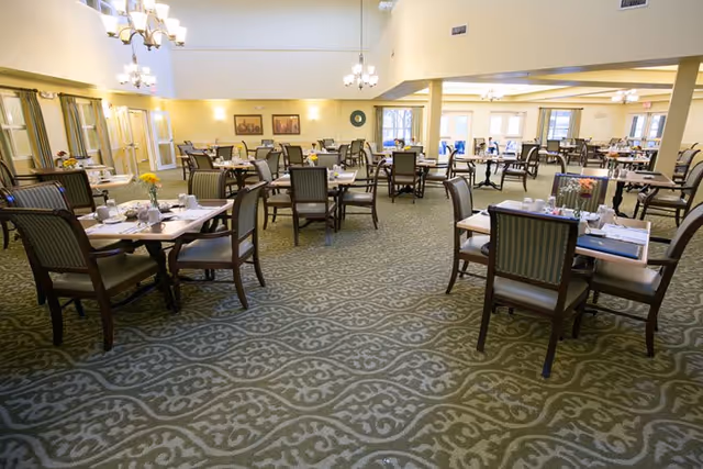 A spacious dining room in a senior living facility with multiple tables and chairs arranged neatly. Each table is set with plates, cups, and napkins, and some have small flower vases. The room has patterned carpet flooring, light yellow walls, and several chandeliers hanging from the ceiling. Large windows allow natural light to brighten the space.