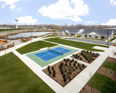 Outdoor view of a senior living facility featuring a blue and green pickleball court surrounded by sidewalks, landscaped areas with shrubs, a pond with a walking path, and single-story residential buildings under a partly cloudy sky.