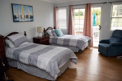 A bright bedroom with two twin beds featuring gray and white striped bedding. Between the beds is a wooden nightstand with a lamp. A painting of sailboats hangs on the wall above one bed. The room has wooden flooring, a blue recliner chair, and large windows with pink curtains letting in natural light. A glass door leads to an outdoor patio area with a table and umbrella visible outside.