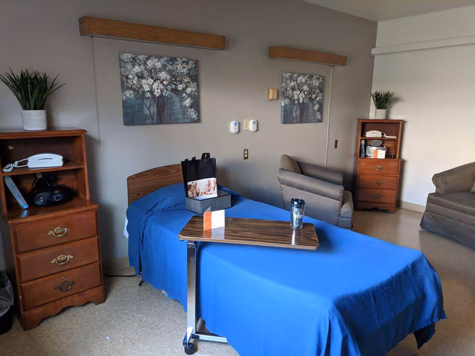A senior living facility bedroom with a single bed covered in a blue blanket. There is a wooden bedside table with a white telephone and a plant on top, and a similar wooden cabinet on the opposite side with another plant and some items. Two framed floral paintings hang on the wall above the bed. A small overbed table with a tumbler and a gift bag is positioned over the bed. Two gray armchairs are placed near the wall.