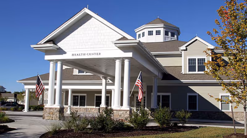 Front entrance of a health center building with a covered portico, white columns, American flags, and landscaped grounds.