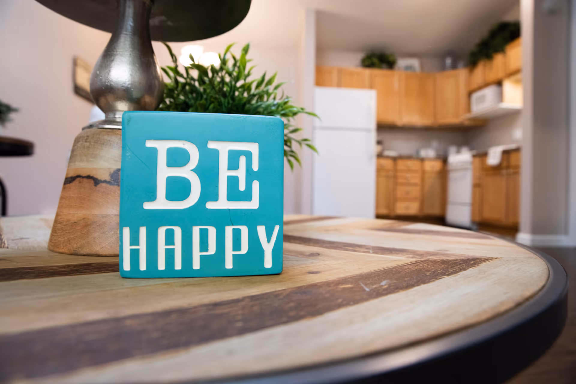A teal decorative block reading "BE HAPPY" sits on a wooden table beside a lamp and plant with a kitchen visible in the background.
